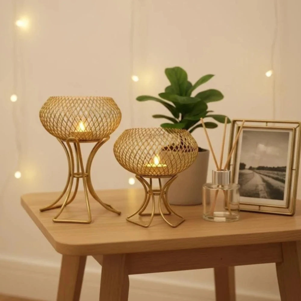 Two gold-toned metal candle holders with woven spherical tops and candle flames inside, displayed on a light wood table alongside a green plant, a framed black-and-white photo, and a glass reed diffuser against a softly lit wall with string lights.
