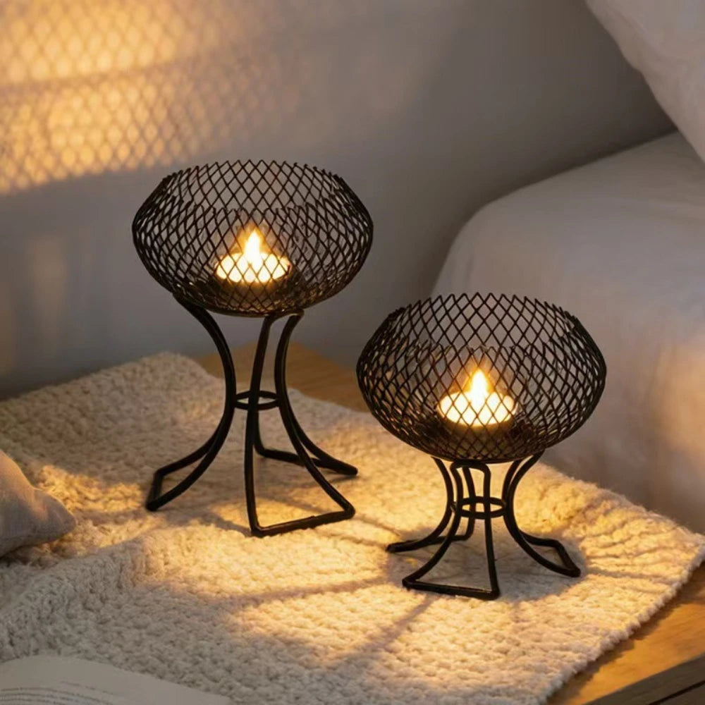 Two black metal mesh candle holders with candle flames lit, placed on a textured cream blanket in a softly lit bedroom setting.