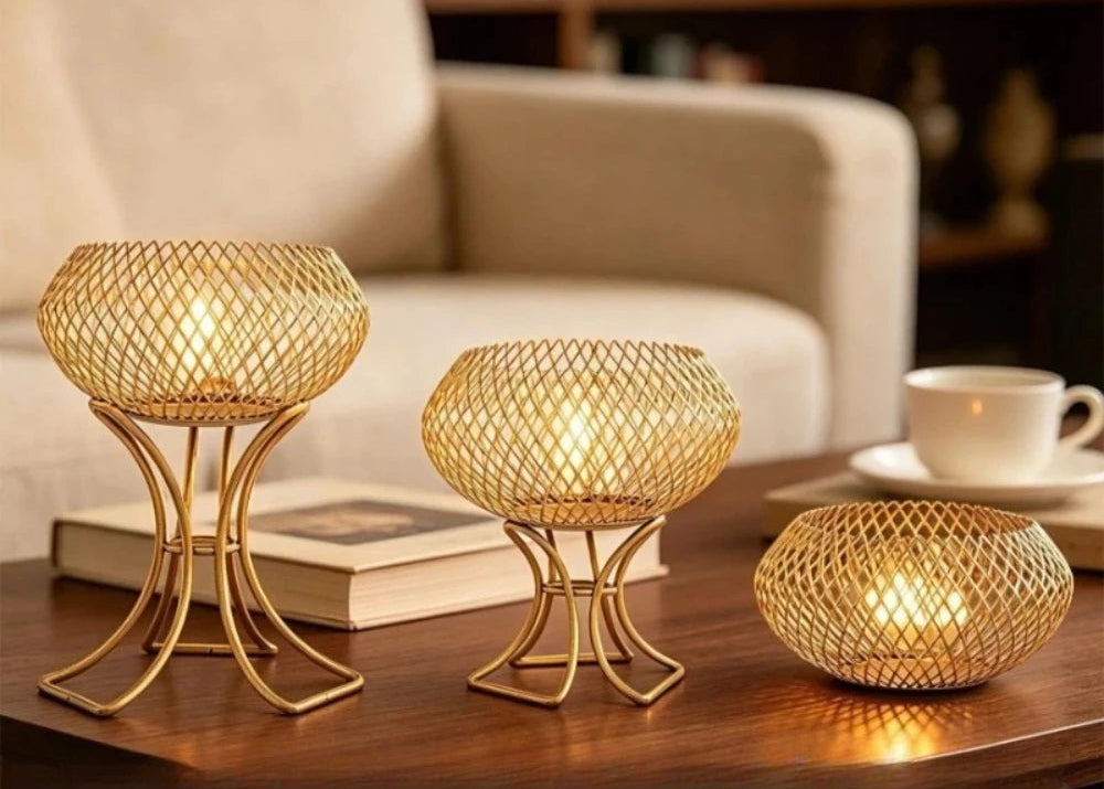 Three gold-toned metal candle holders with woven mesh bowls and curved pedestal stands are shown on a wooden table in a living-room setting, with a sofa, book, and cup visible. A size comparison chart appears along the bottom showing different sizes and weights of the same holder.