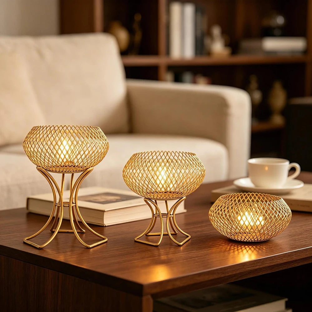 Three gold-toned metal mesh candle holders with wireframe bowls and curved stands, each holding a warm lit candle, arranged on a wooden table in a cozy living room setting.