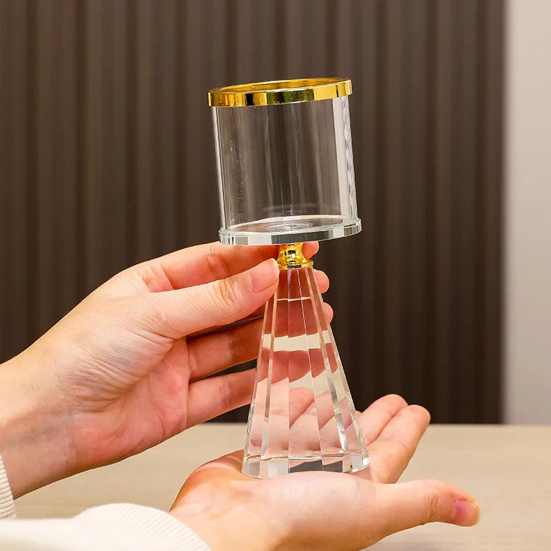 A clear glass hourglass with a gold-colored rim and a faceted clear glass base, held by two hands against a dark brown paneled background.