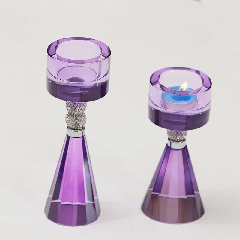 Two purple glass candle holders with faceted cone bases and crystal-like decorative necks, shown on a plain light background. One holder is empty, and the other holds a small blue tea light candle.