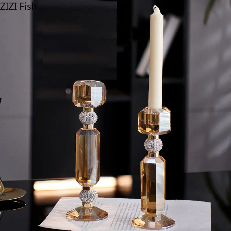 Two elegant gold-toned crystal candlestick holders with a tall white taper candle, displayed on a dark reflective surface with a blurred indoor background.