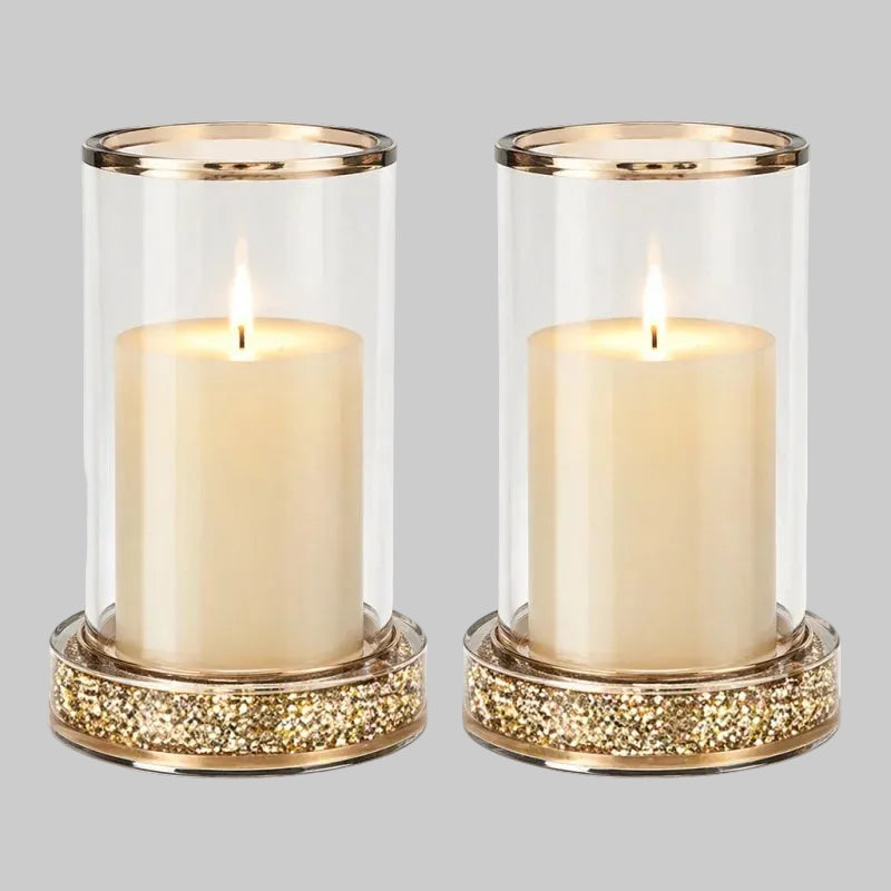 Two matching clear glass candle holders with gold trim, each containing a lit cream pillar candle. The holders have a wide, circular base covered in shimmering gold glitter, set against a plain white background.