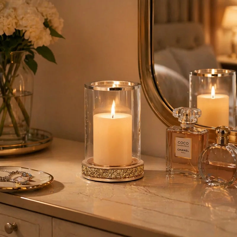A lit white pillar candle in a clear glass cylinder holder with a gold-toned decorative base, styled on a vanity table beside perfume bottles and a mirror.