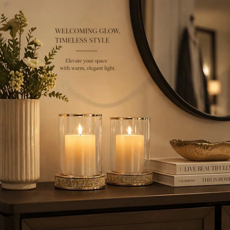 Two lit glass candle holders with gold trim and glittery bases displayed on a dark wooden console table in a warmly lit room. A vase with white flowers, a round mirror, and stacked books are visible in the background, with decorative text on the wall reading 'WELCOMING GLOW, TIMELESS STYLE'.