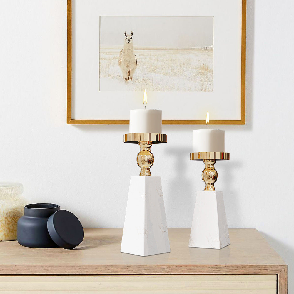 Two white marble candlestick holders with gold metal accents, each holding a white pillar candle, styled on a light wood console table against a white wall with a framed picture.
