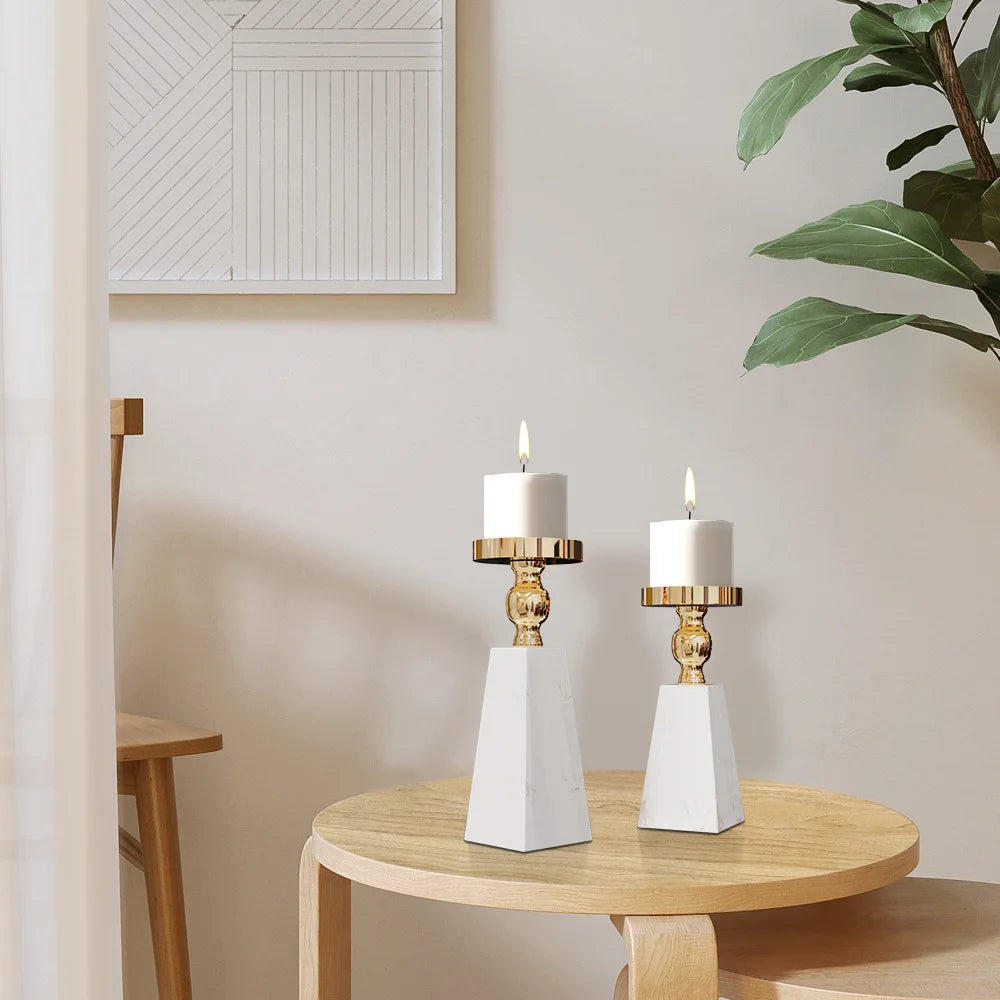 Two white candle holders on a wooden table with a plant and framed picture in the background.