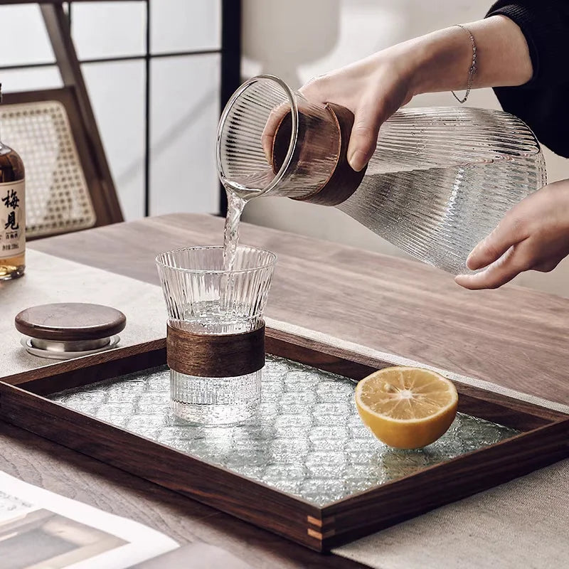 Person pouring liquid from a glass container into a small cup on a wooden tray with a lemon half.