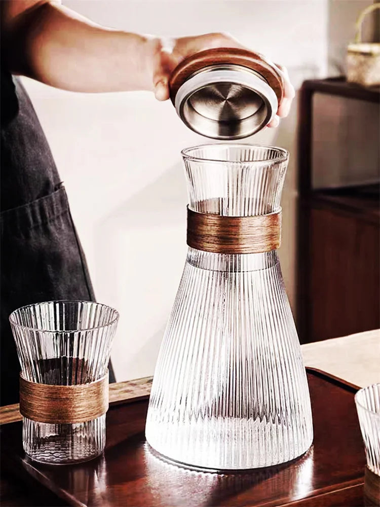 Ribbed glass carafe with wooden accents on a wooden surface, with a person holding a lid.