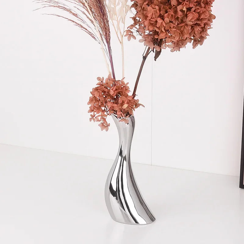 A modern, curved metallic silver vase filled with dried orange-brown flowers and pampas grass, set against a clean white background.