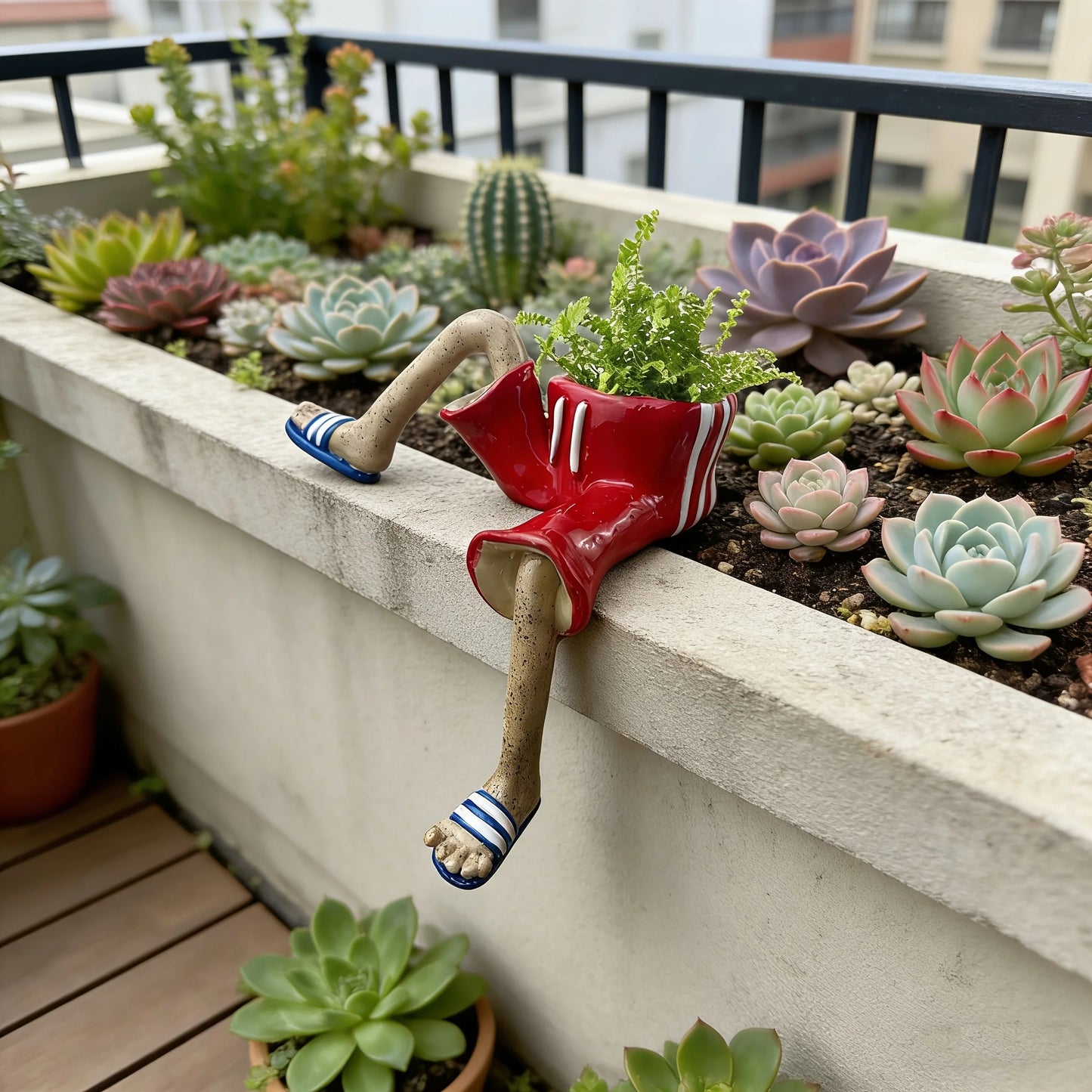 A red planter with a dangling leg and flip-flop foot, filled with green succulents and small plants, displayed in a concrete planter box on a balcony with other potted succulents.