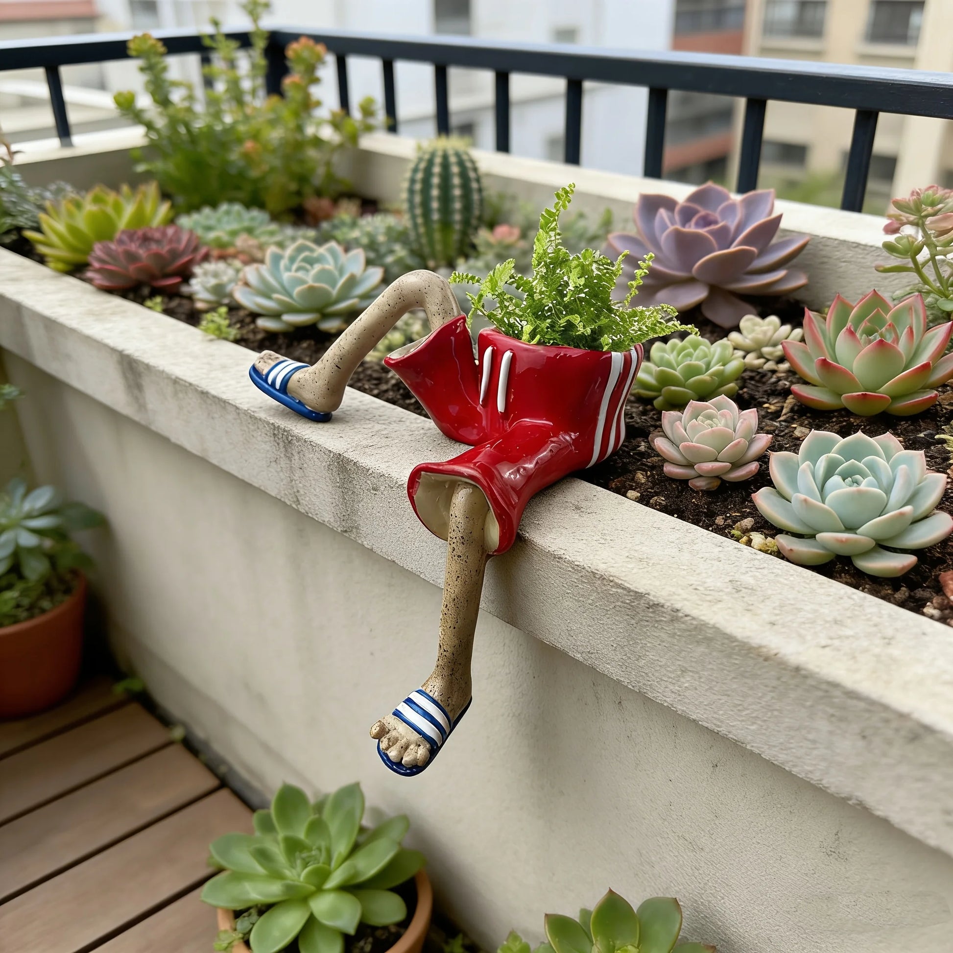 A red planter with a dangling leg and flip-flop foot, filled with green succulents and small plants, displayed in a concrete planter box on a balcony with other potted succulents.