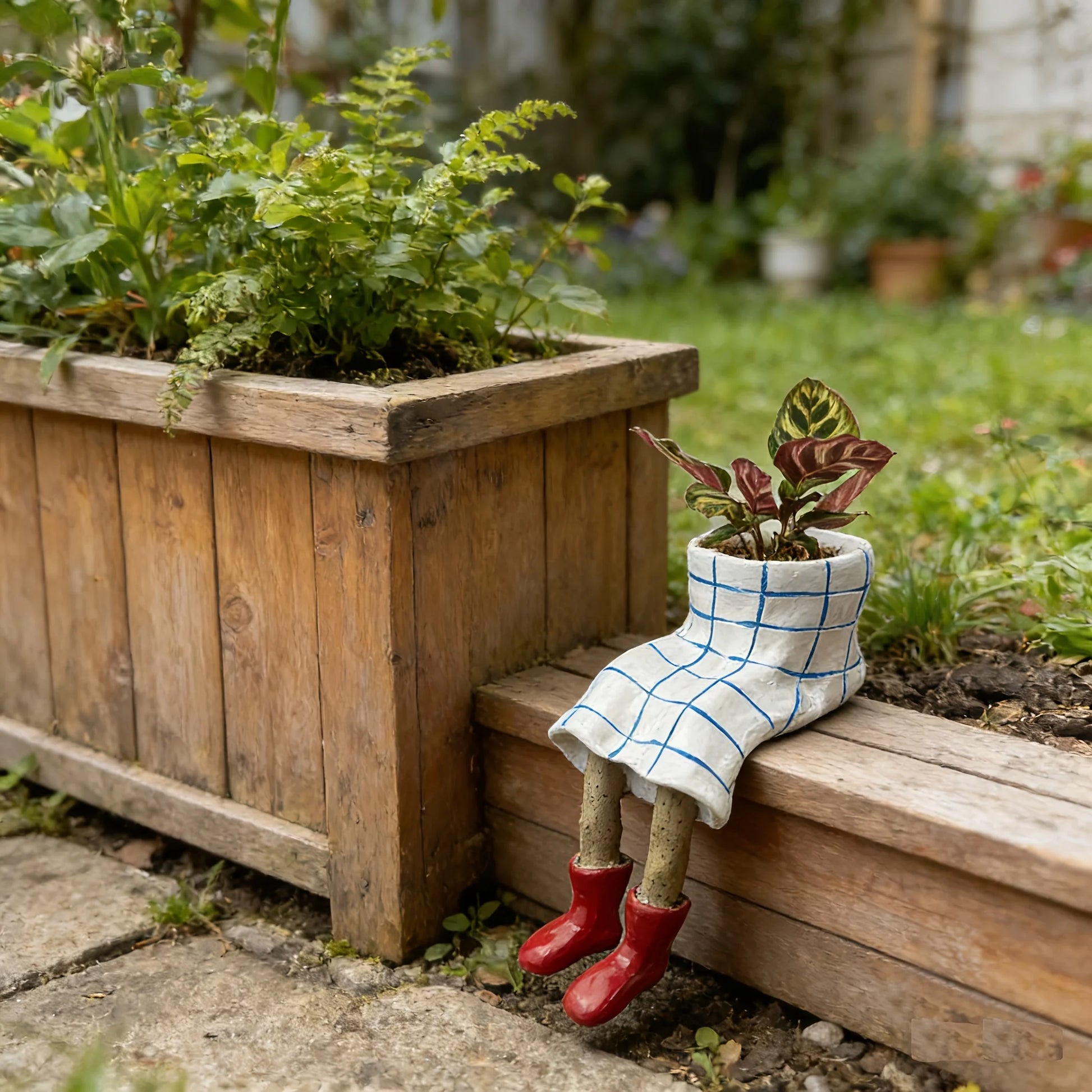Small garden pot in a plaid outfit and red boots sitting on a wooden planter with plants.