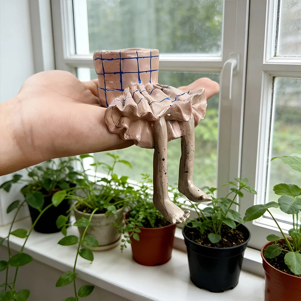 A handmade ceramic miniature figurine of a seated figure with a plaid skirt, held in a person's hand in front of a window with potted plants.