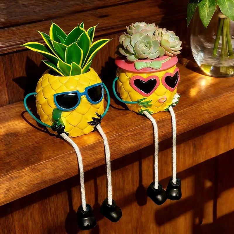 Two decorative pineapple-shaped planters with faces, sunglasses, and dangling legs, one yellow with blue sunglasses and a green succulent, the other yellow with pink heart-shaped sunglasses and a pink hat, both sitting on a wooden shelf.