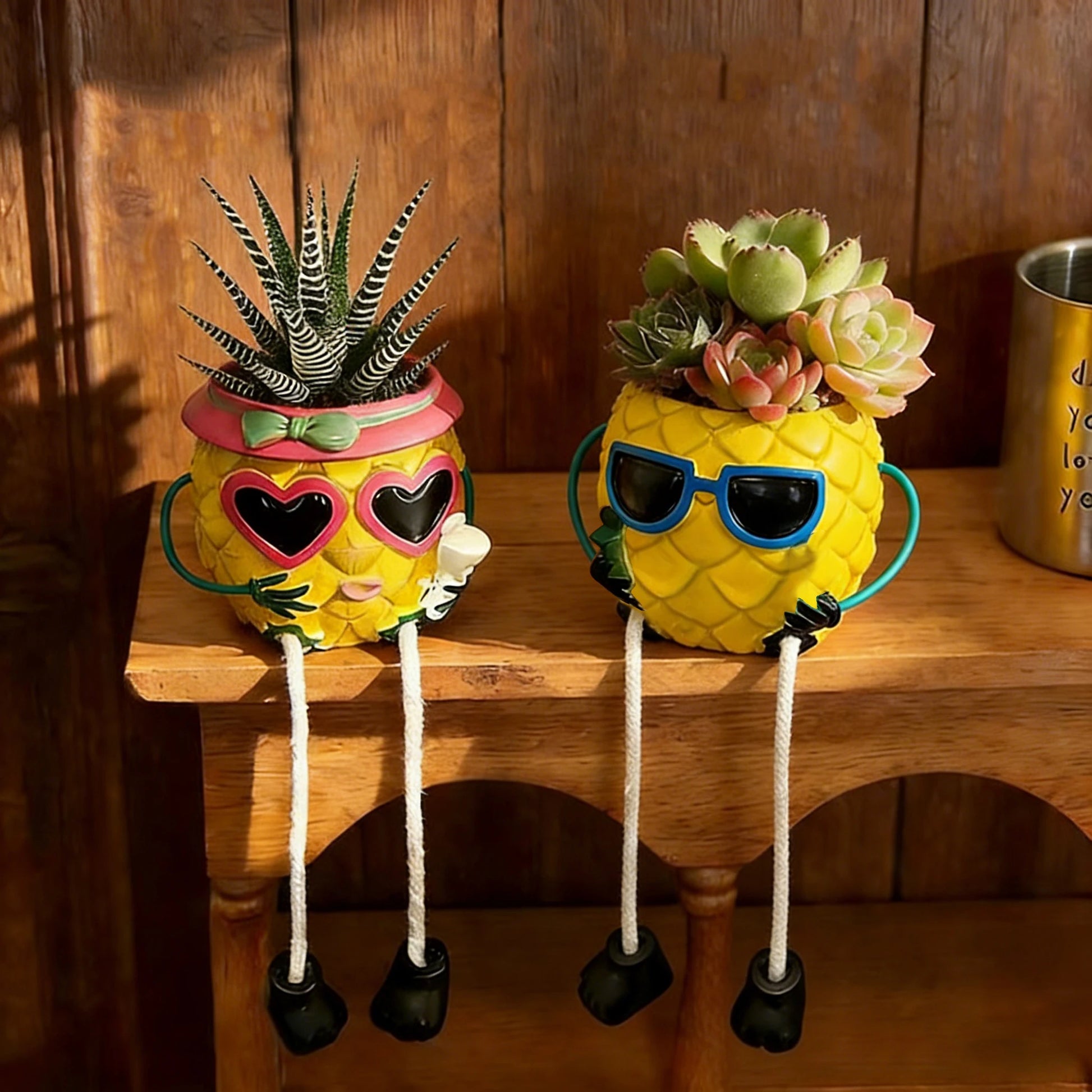 Two pineapple-shaped planter pots with legs and sunglasses, sitting on a wooden shelf. The left planter has a pink heart-shaped rim and a small succulent, while the right planter has blue sunglasses and a larger green succulent.
