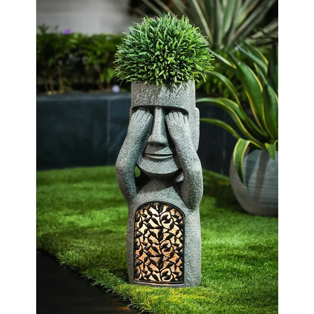 Decorative stone statue - see no evil - with face and plant on head, placed outdoors on grass.