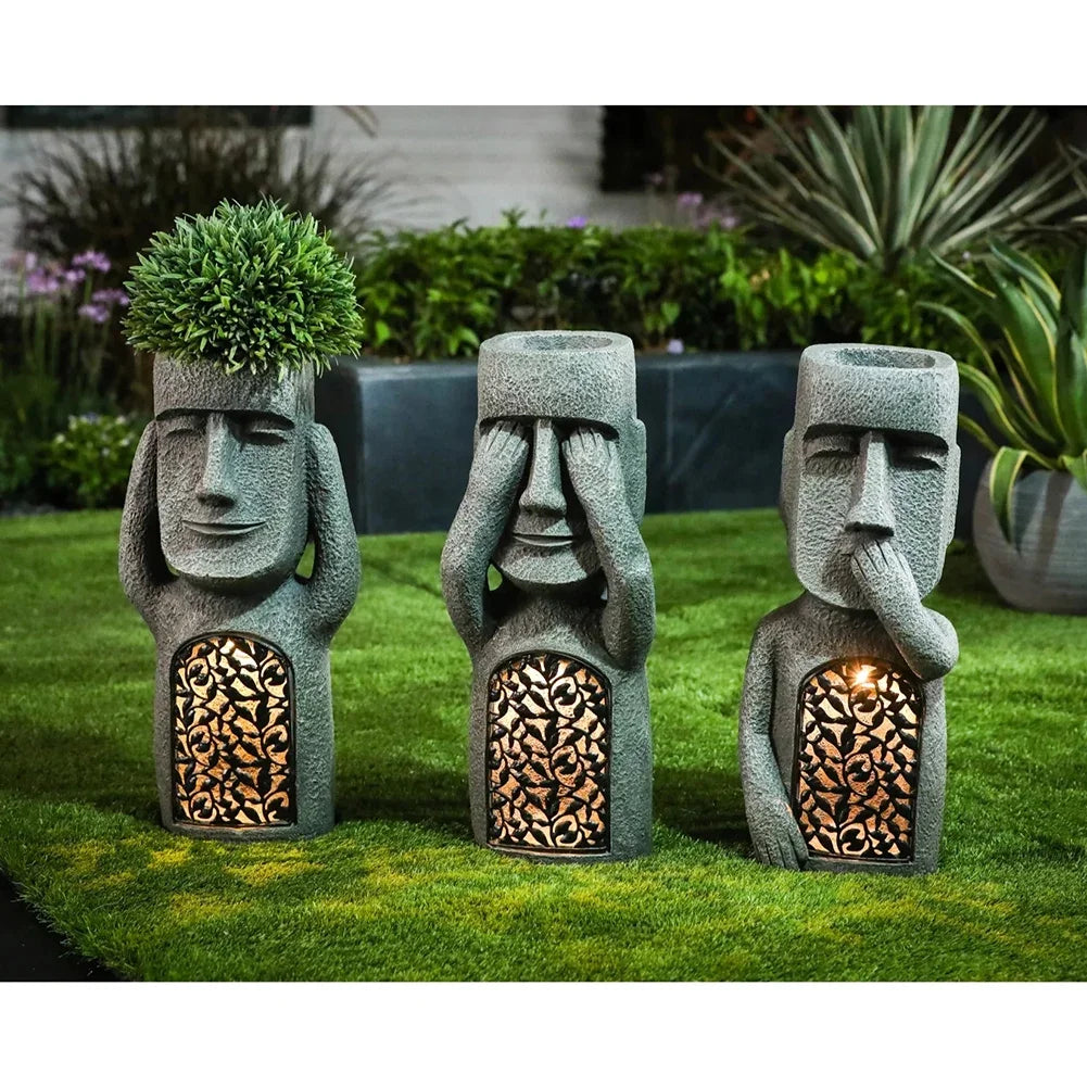 Three stone-like statues with faceless designs, each with a different pose, on grass with plants in the background.