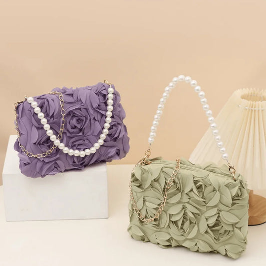 Two textured fabric evening bags with 3D floral rosette and petal designs, featuring pearl-embellished chain straps, displayed on a beige background with one bag on a white block.