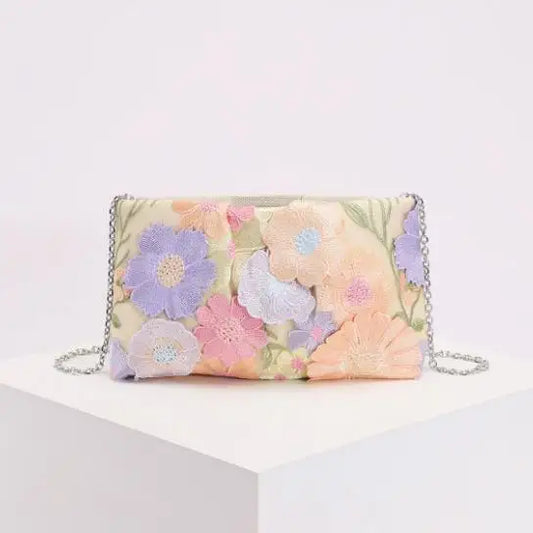 A rectangular clutch bag with a pastel floral patchwork design featuring embroidered flowers in pink, purple, peach, and blue on a cream background, with a silver chain strap, displayed on a white block against a white background.