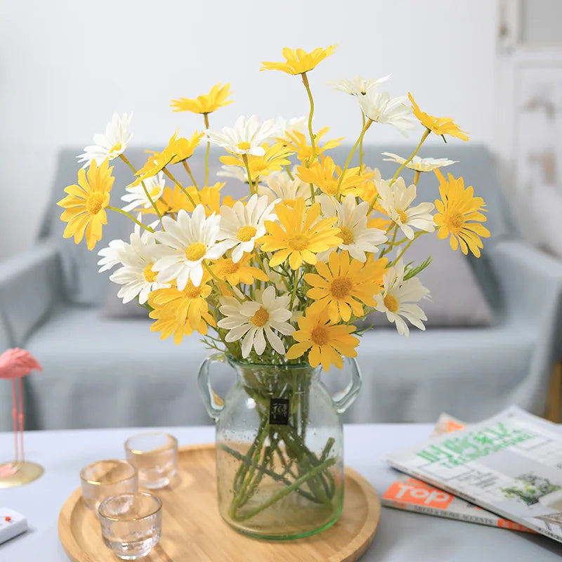 Bouquet of yellow and white flowers in a clear vase on a table with a light blue sofa in the background.