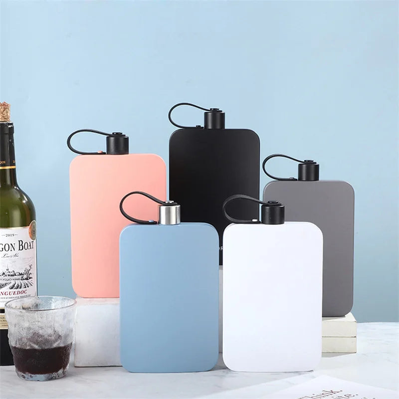 Five rectangular hip flasks in pastel pink, black, light blue, white, and dark gray, arranged on stacked white blocks against a light blue background. A wine bottle and a glass of dark wine are partially visible on the left.