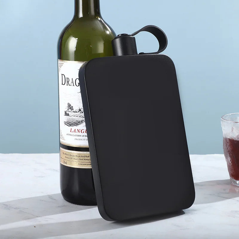 A matte black rectangular flask with a matching black screw-top lid and loop handle, shown leaning against a wine bottle on a light marble surface. A glass of red wine is partially visible at the right edge.