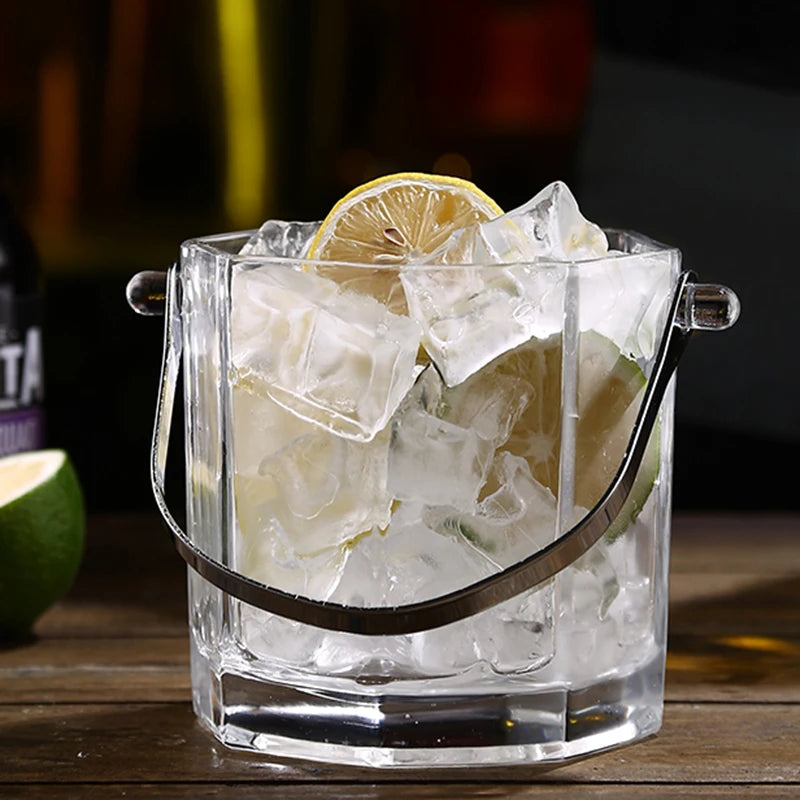 Clear octagonal ice bucket with a metal handle, filled with large ice cubes and garnished with a lemon slice, set on a wooden surface.