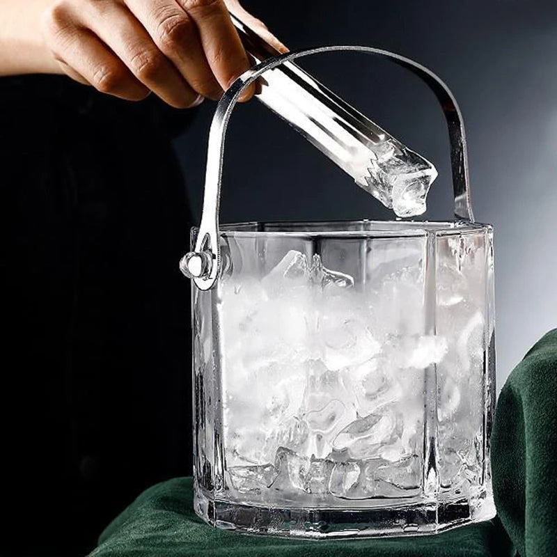 A clear glass ice bucket filled with ice, shown with a hand placing a glass ice scoop into it against a dark background.