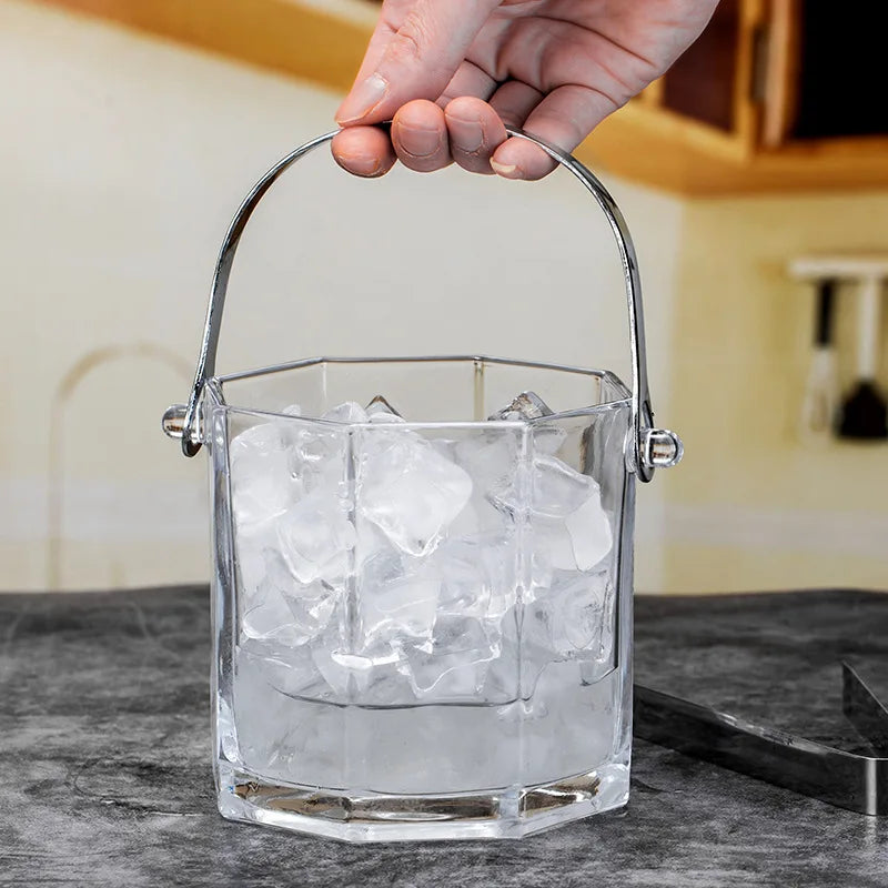 A clear glass bucket-style ice bucket filled with ice cubes, held by a hand with a metal handle, set on a dark countertop in a kitchen environment.