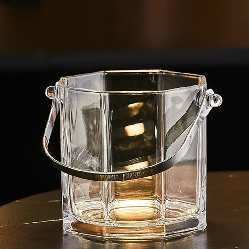 A clear glass bucket-style container with a hexagonal base, ribbed sides, and a curved metal handle, shown on a dark tabletop against a dark background.