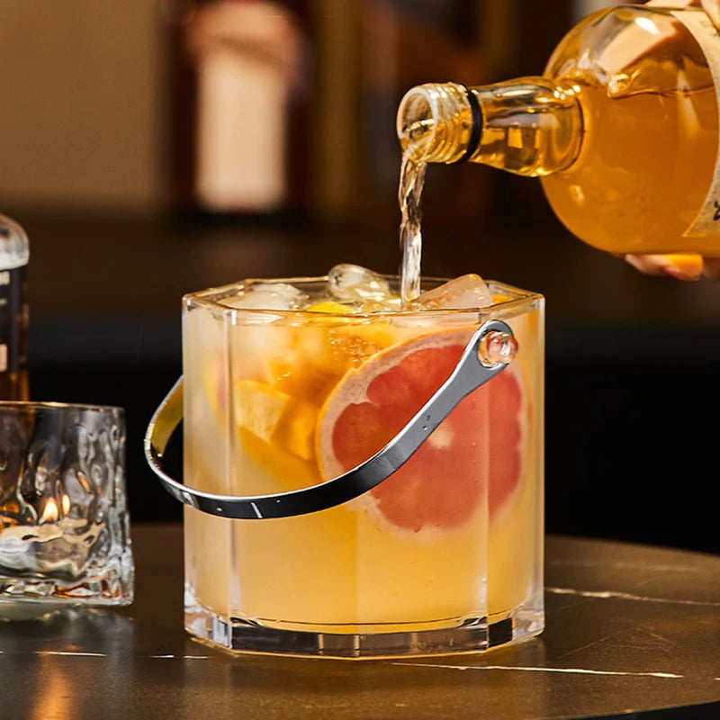 A clear ice bucket cocktail glass with a metal handle, filled with an amber drink and garnished with a grapefruit slice, while a bottle pours the liquid into the glass on a bar counter.