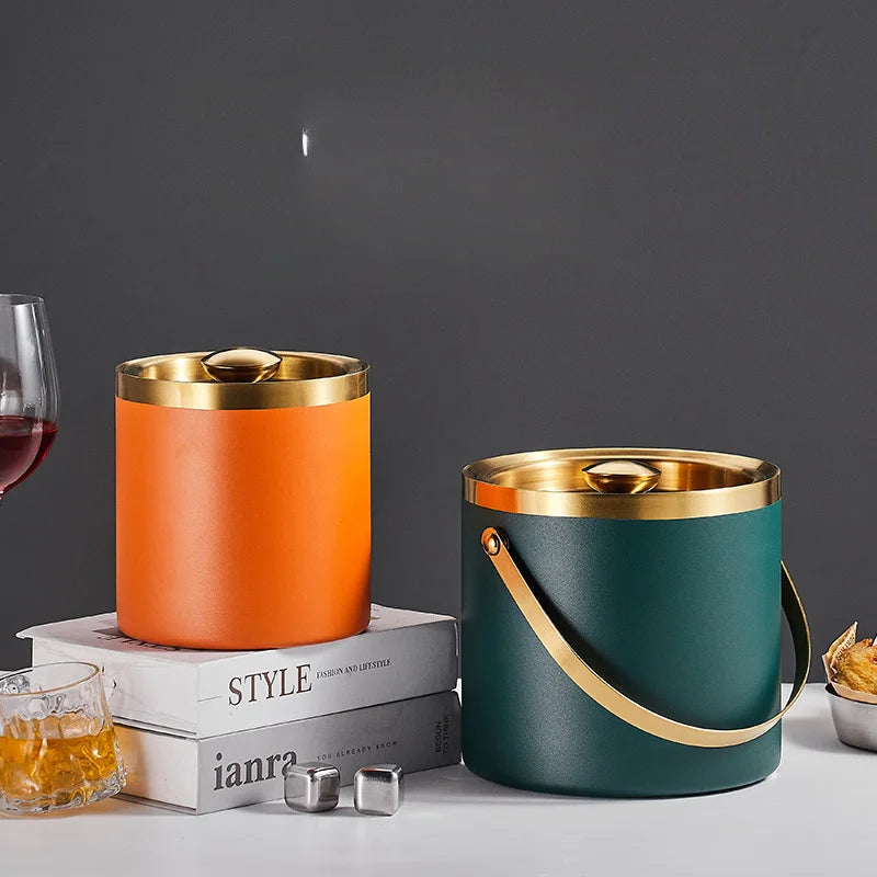 Two insulated bucket-style ice buckets with lids are shown on a tabletop against a dark gray background. One bucket is matte orange and the other is matte dark teal/green, both with metallic gold trim and handles.