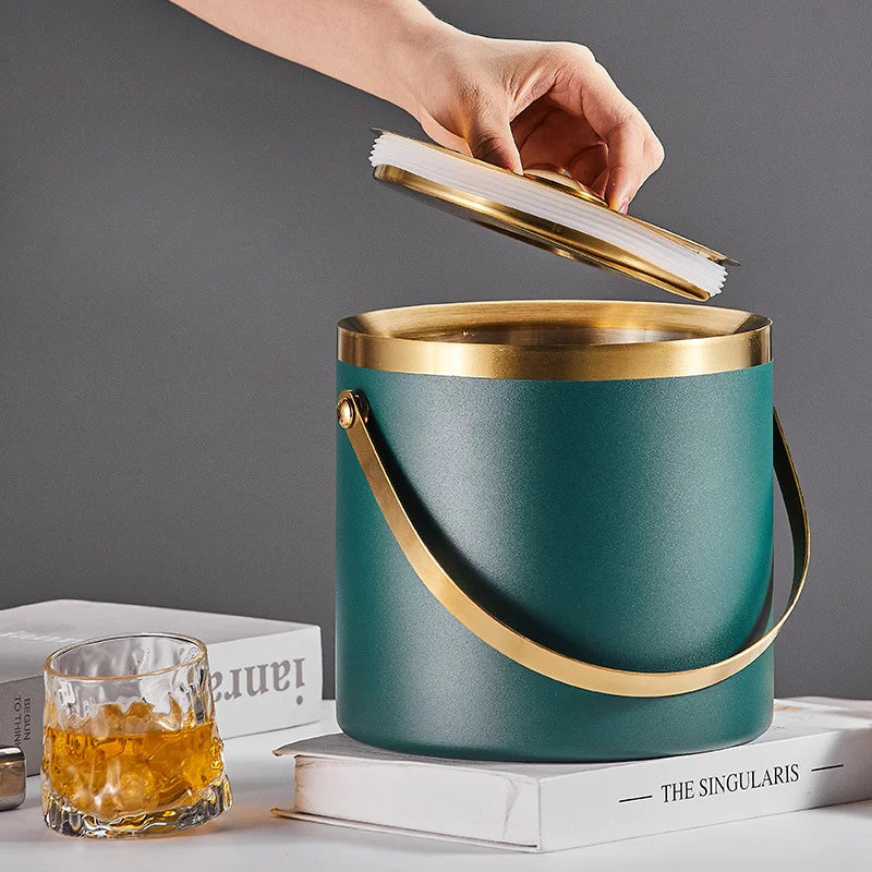 A teal insulated ice bucket with a gold-tone handle and lid, shown with a hand lifting the lid. It is styled on white books with a glass of amber liquid nearby against a dark gray background.