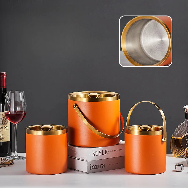 Three orange insulated drink buckets with gold-tone metal rims and handles, displayed on a tabletop with a wine bottle, wine glass, books, and a perfume bottle. An inset close-up at the top right shows the brushed stainless steel interior of the largest bucket.