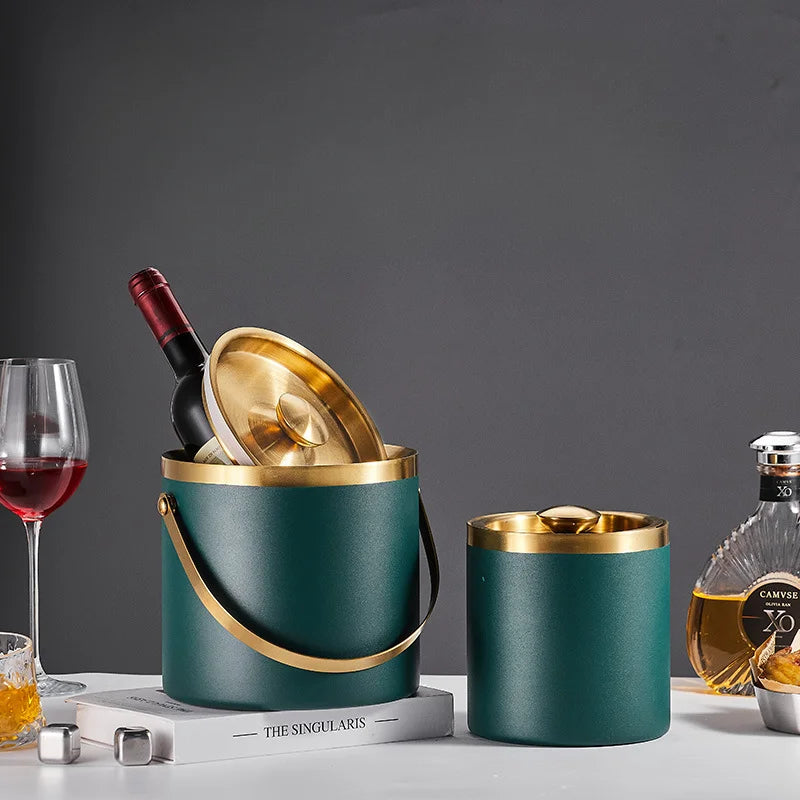 A dark teal ice bucket with gold trim and handle, shown with a wine bottle inside and a matching lid beside it on a tabletop. A wine glass, book, and liquor bottle are styled around it against a dark gray background.