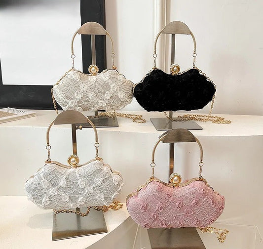 Four small evening handbags displayed on metal stands in a showroom. The bags have scalloped floral lace or velvet exteriors in white, black, and pink, with gold-tone handles, chain straps, and oval clasps.