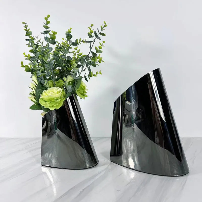 Two modern glossy black cone-shaped decorative vases on a white marble surface against a white wall, with one vase holding green artificial foliage.