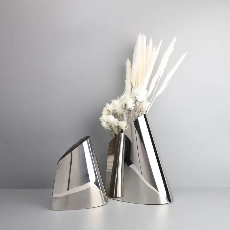 Two modern metallic cone-shaped vases in polished silver, one empty and one holding dried white pampas grass, set against a neutral gray background.