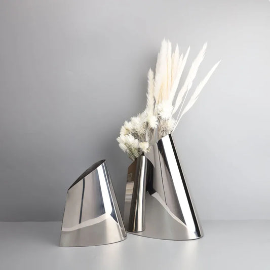 Two modern metallic cone-shaped vases in polished silver, one empty and one holding dried white pampas grass, set against a neutral gray background.