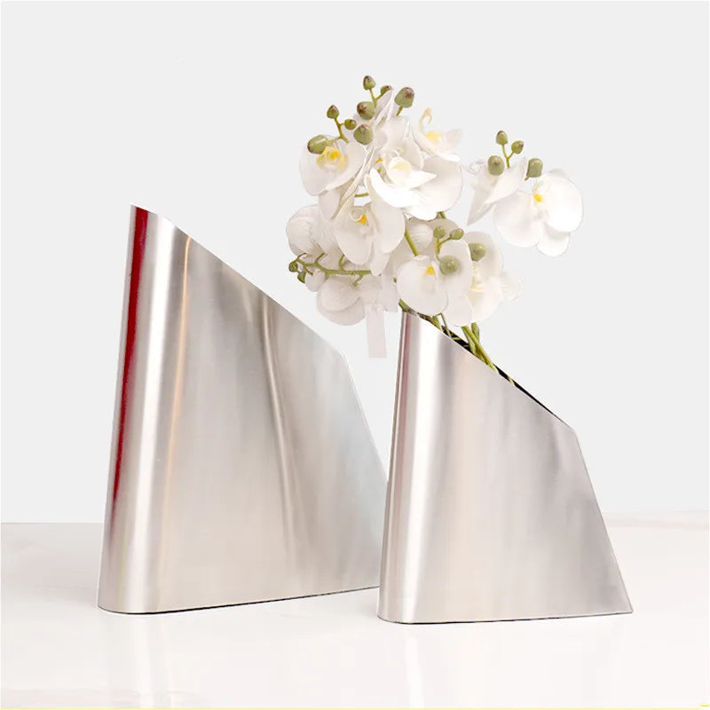 Two modern metallic silver geometric flower vases on a white surface against a light gray background, with white orchids placed in the right vase.