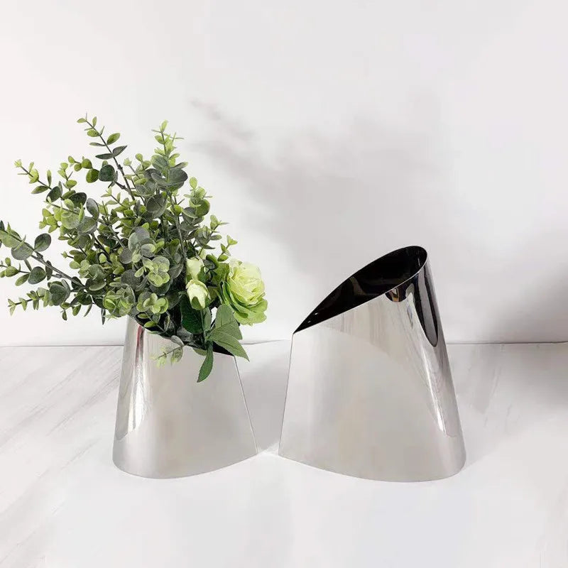 Two modern metallic cone-shaped planters with a reflective silver finish, one holding green artificial foliage and the other empty, set against a clean white background.