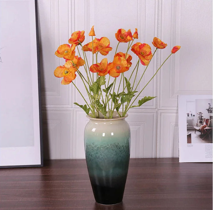 Decorative vase with orange flowers on a wooden floor against a white paneled wall.
