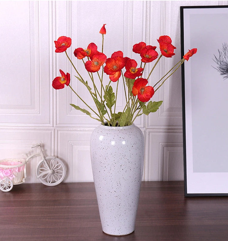 White vase with red flowers on a wooden surface against a white paneled wall.