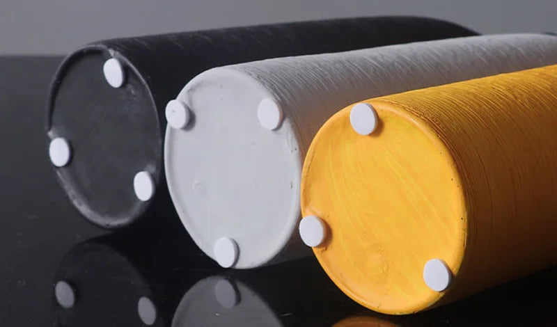 Three cylindrical vases in grey, white, and yellow, each with four small white circular studs or pads on the end, arranged on a reflective black surface.