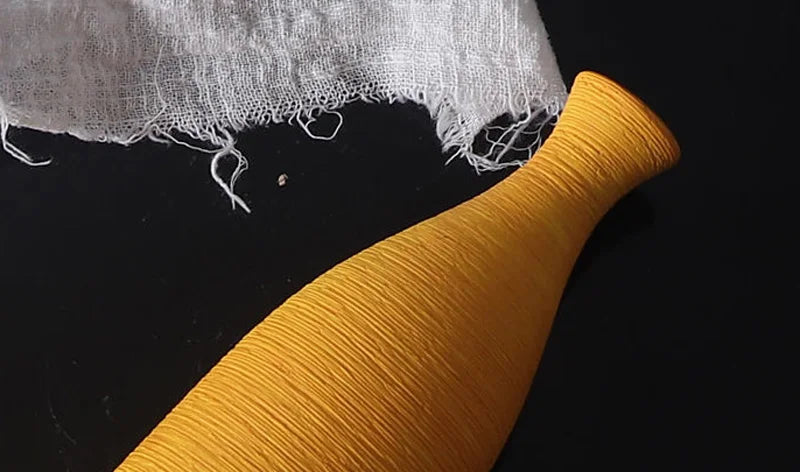 A mustard yellow textured bottle-shaped vase or decorative object shown on a black background with a piece of white gauze fabric at the top.