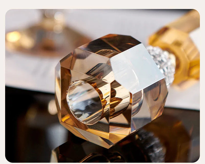A close-up of a faceted crystal ring with a gold-toned band and a clear rhinestone accent, shown on a dark reflective surface with a blurred background.