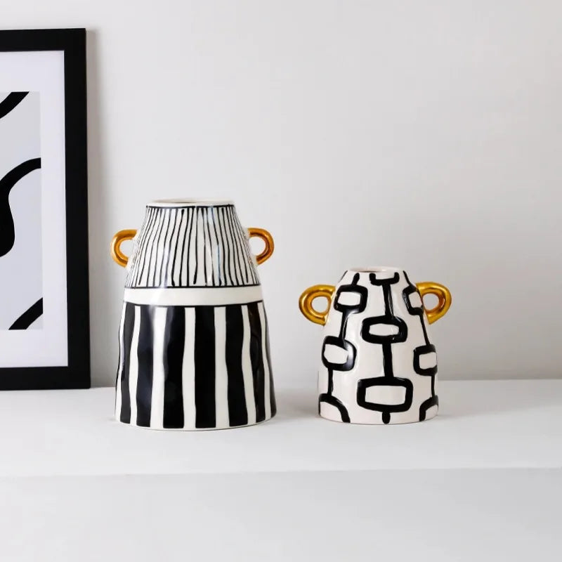 Two decorative ceramic vases with black and white patterns and gold handles, displayed on a white shelf next to a framed print against a white wall.