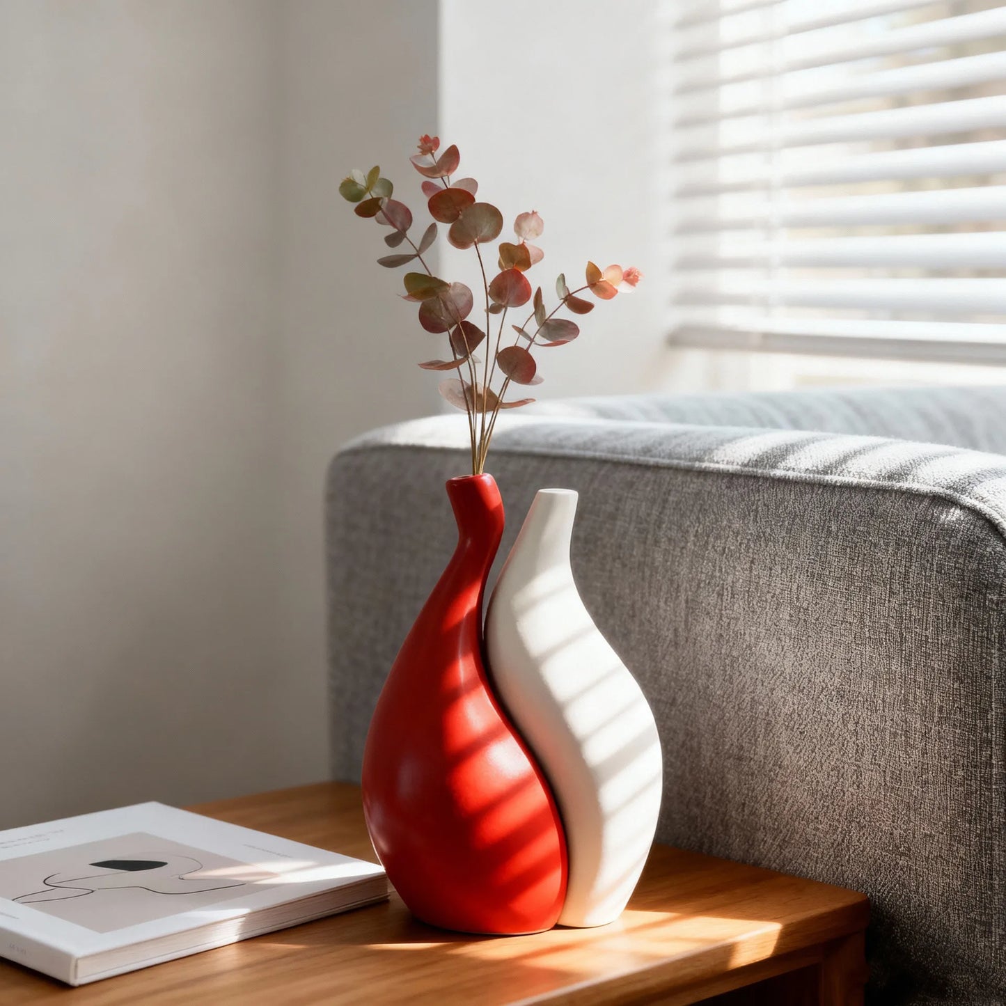 Two modern ceramic vases, one glossy red and one matte white, arranged side by side on a wooden table next to a gray sofa with eucalyptus branches in the red vase.